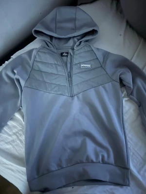 Grå hoodie från McKenzie med dragkedja - Snygg grå hoodie från McKenzie med halv dragkedja framtill och quiltad detalj över bröstet. Hoodien har en klassisk huva och ribbade muddar vid ärmslut och nederkant. Perfekt för en chill och sportig stil.