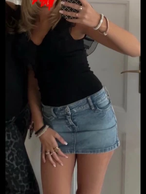 Svart body med volangdetaljer - Snygg svart body med djup v-ringning och volangdetaljer i mesh längs axlar och halsringning. Tight passform och stretchigt material som sitter bekvämt. Perfekt att styla med kjol eller jeans för en trendig look.
