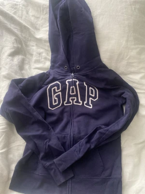 Mörkblå hoodie från GAP med dragkedja - Mörkblå hoodie från GAP med vit broderad logga på bröstet. Tröjan har huva med dragsko, dragkedja framtill och två fickor. Perfekt för en avslappnad och cool stil. Tillverkad i mjukt bomullsmaterial.