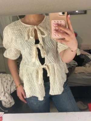 Vit strukturerad blus med knyt - Superfin vit blus med strukturmönster och tre knyt framtill. Blusen har korta puffärmar och en lös, flowy passform. Perfekt att styla med jeans eller kjol. Använd få gånger, vit/cremefärgad