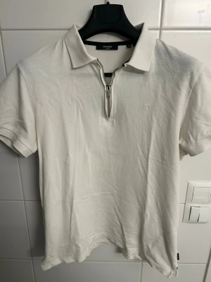 Vit polotopp med dragkedja Jack & Jones - Vit polotopp från Jack & Jones med kort ärm och klassisk krage. Toppen har en snygg dragkedja framtill och är tillverkad i ett mjukt bomullsmaterial. Perfekt för en clean och stilren look.