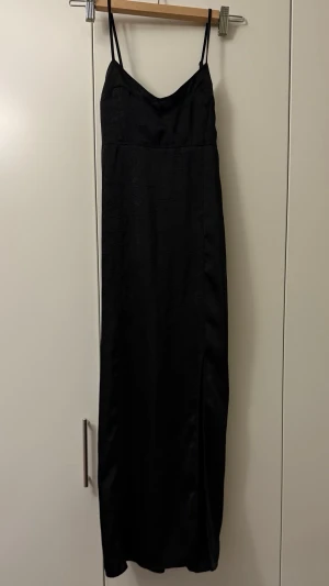 Svart långklänning med slits - Svart långklänning med smala axelband och v-ringad hals. Klänningen har en elegant slits på sidan och är gjord i ett glansigt, lätt material som ger en snygg siluett. Perfekt för dig som gillar stilrena och klassiska plagg.