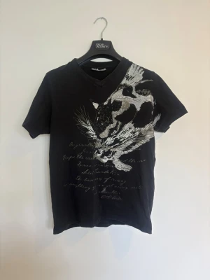 Svart t-shirt med fågeltryck - Cool svart t-shirt från OAK Jeans med v-ringning och korta ärmar. Framsidan har ett stort tryck av en fågel i vitt och grått samt textdetaljer. Perfekt för en avslappnad och trendig look.