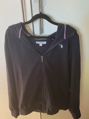 Marinblå hoodie från U.S. Polo Assn - Mörkblå hoodie med dragkedja från U.S. Polo Assn. Klassisk design med vit logga på bröstet och huva med rödvit randig detalj på insidan. Mjuk bomullskänsla och ribbade muddar vid ärmslut och nederkant. Perfekt för en avslappnad stil.