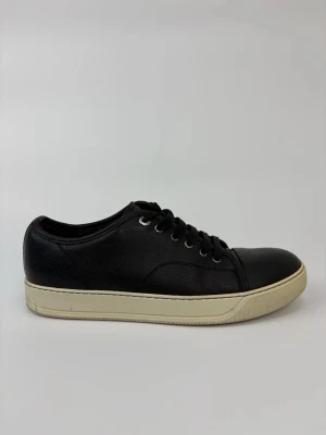 Svarta Lanvin sneakers med vit sula - Stilrena svarta sneakers med vit sula och svarta skosnören. Skorna har en klassisk låg profil och är tillverkade i slätt och lätt texturerat material. Metallfärgade öljetter ger en snygg detalj. Perfekta för en clean och enkel look.