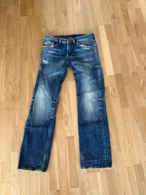 Blå slitna jeans från Diesel - Säljer ett par blå jeans från Diesel med snygga slitningar och distressed detaljer. Jeansen har raka ben, klassisk femficksmodell och tydliga slitningar på låren och knäna. Perfekt för dig som gillar en avslappnad och trendig stil.