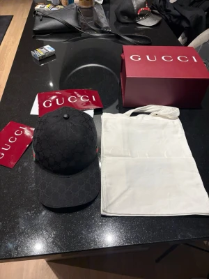Svart keps från Gucci med monogram - Snygg svart keps från Gucci med klassiskt GG-monogram över hela kepsen. På sidan finns det ikoniska gröna och röda bandet. Kepsen har böjd skärm och justerbar passform. Perfekt accessoar för att lyfta din streetwear-look.