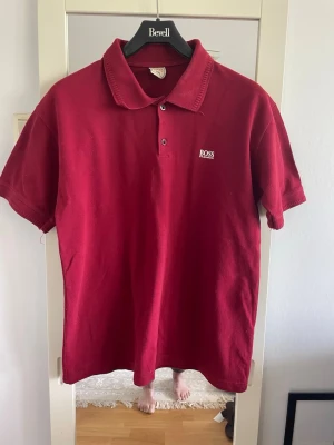 Vinröd pikétröja från BOSS - Vinröd pikétröja från BOSS med klassisk krage och två knappar framtill. Tröjan har korta ärmar och en broderad BOSS-logga på bröstet. Perfekt för en stilren och avslappnad look.