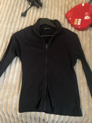 Svart ziptröja från Gina Tricot - Säljer en svart ziptröja från Gina Tricot med hög krage och hel dragkedja framtill. Tröjan har en enkel och stilren design med långa ärmar och är perfekt för lager på lager. Materialet känns mjukt och stretchigt.