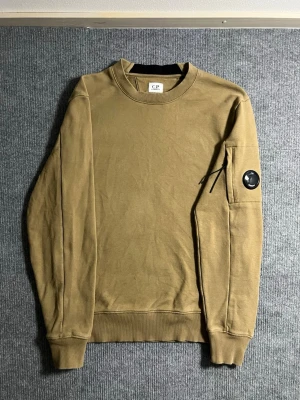 C.P. Company – Khaki Lens Sweatshirt (S) - Stylish and iconic C.P. Company sweatshirt in a classic khaki shade. Size S, length 64 cm, width 49 cm – fits perfectly for a modern, relaxed look. The sleeve features the signature lens detail, instantly recognizable to fans of the brand.  ✅ Condition: Excellent – no wear, no stains 💯 Authenticity: 100% genuine, easily verified on certilogo.com 📦 Shipping: Sent immediately after purchase – fast and effortless transaction!  This sweatshirt combines C.P. Company’s technical style with minimalist 