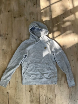 Grå hoodie från Lyle & Scott - Snygg grå hoodie från Lyle & Scott med klassisk gul logga på bröstet. Hoodien har huva med dragsko, magficka och ribbade muddar vid ärmar och nederkant. Tillverkad i mjuk bomullsblandning som är skön att ha på sig.