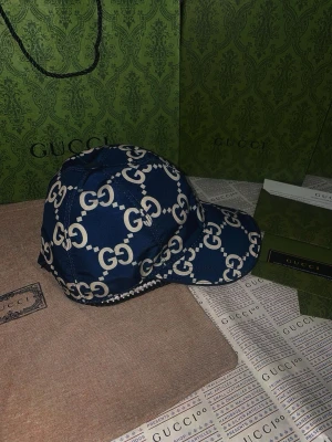 Blå Gucci keps med GG-mönster - Snygg blå keps från Gucci med det klassiska vita GG-monogrammet över hela kepsen. Kepsen har böjd skärm och justerbar passform. Tillverkad i ett glansigt tyg som ger en lyxig känsla. Perfekt för dig som vill sticka ut med en exklusiv accessoar.