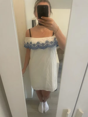 Vit offshoulder klänning med blå spets - Söt vit kortklänning med offshoulder-modell och blå broderad spetsdetalj längs överdelen. Klänningen har en rak passform och är perfekt för varma dagar. 