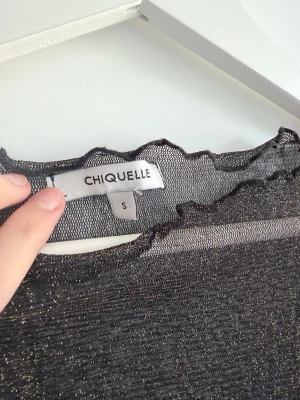 Glittrig svart festtopp från Chiquelle - Snygg svart långärmad festtopp från Chiquelle med glittrigt material och transparent känsla. Toppen har vågiga kanter vid hals, ärmslut och nederkant som ger en extra detalj. Jättefin med spetsbh eller croptop under🤍.