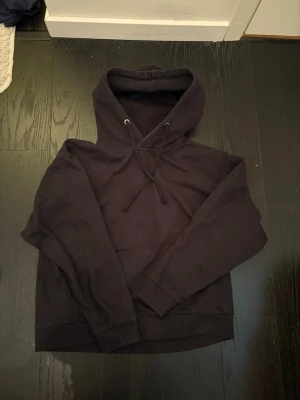 Svart hoodie från 157 - En klassisk svart hoodie från 157 med huva och dragsnören. Hoodien har långa ärmar och ribbade muddar vid ärmslut och nederkant. Tillverkad i mjukt bomullsmaterial som är skönt att bära. Perfekt för en avslappnad och stilren look.