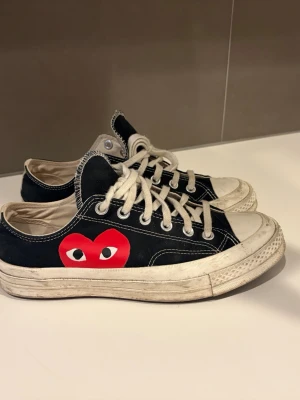 Converse x CDG Play svarta sneakers - Säljer ett par Converse x Comme des Garçons Play sneakers i svart canvas med vit sula och vita snören. Skorna har det ikoniska röda hjärtat med ögon på sidan och klassisk låg modell. Perfekta för dig som gillar streetwear och vill sticka ut.