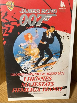 James Bond 007: I Hennes Majestäts Hemliga Tjänst VHS - Säljer en svensktextad VHS med James Bond-filmen 'I Hennes Majestäts Hemliga Tjänst' med George Lazenby som agent 007. Omslaget är rött, svart och vitt med klassisk Bond-design och actionmotiv. Perfekt för samlare av Bond eller retrofilmer.