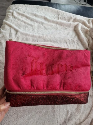 Rosa necessär från Juicy Couture - Säljer en snygg rosa necessär från Juicy Couture med glittrande röda detaljer och dragkedja. Mjuk sammetliknande yta och texten 'Juicy' i röda stenar framtill. Perfekt för smink eller småprylar.