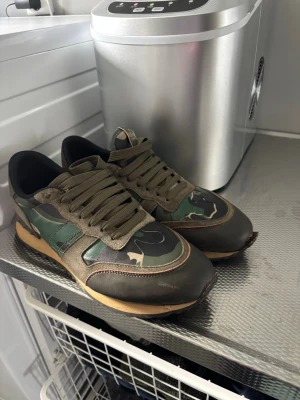  Valentino rockrunners  - Snygga sneakers från Valentino med grön och brun camo-design, svarta och olivgröna detaljer samt beige sula. Skorna har klassisk låg profil, gröna skosnören och paneler i mocka och läder. Perfekta för dig som vill sticka ut med streetwear-stil. Strl 46 
