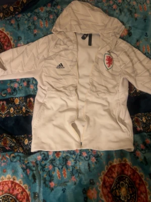 Vit Adidas hoodie med Wales-emblem - Snygg vit hoodie från Adidas med grått mönster och Wales-emblem på bröstet. Dragkedja framtill, två fickor och klassisk huva. Perfekt för dig som gillar sportig stil och vill sticka ut med ett unikt plagg.