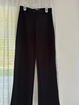 Svarta vida kostymbyxor - Stilrena svarta kostymbyxor med raka, vida ben och klassisk midja. Byxorna har knapp och dragkedja framtill samt hällor för skärp. Perfekta för en clean och dressad look. 