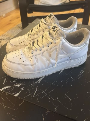 Nike Air Force 1 vita sneakers - Klassiska vita Nike Air Force 1 sneakers i lågt utförande. Skorna har en ikonisk siluett med perforerad tå, tjock sula och Nike-logga på sidan. Tillverkade i läder med textildetaljer och vita skosnören. Perfekta för en clean och tidlös streetstil. Använda i ca 9 månader men vissa slitningar i skorna men som inte märks av vid användning 