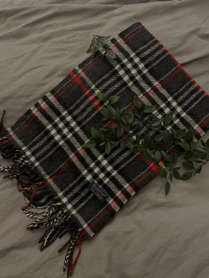 Mörkgrå rutig halsduk i ull - Snygg mörkgrå halsduk från Johnstons of Elgin i mjuk ull. Klassisk rutigt mönster i svart, vitt och rött med tvinnade fransar som avslut. Perfekt accessoar för att lyfta din outfit med en tidlös och stilren.