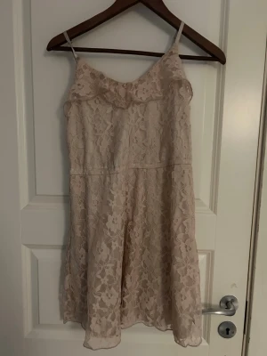 Ljus spetsklänning - Säljer en söt ljus kortklänning från H&M i storlek 34. Klänningen har tunna axelband och är täckt av ett blommigt spetsmönster. Den har en mjuk, beige underton och är fodrad med ett lent tyg. Perfekt för dig som gillar romantisk stil och detaljer.
