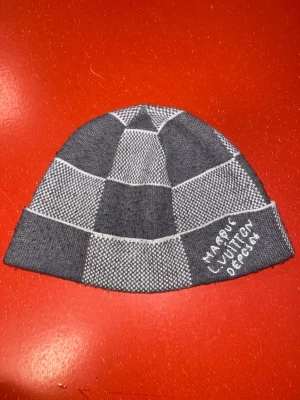 Gråmönstrad mössa  - Snygg grå och vit stickad mössa från med rutigt mönster och texten 'Marque L Vuitton Deposee' broderad på uppviket. Perfekt accessoar för att lyfta din streetwear-look.