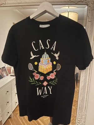 Svart Casa Way t-shirt med tryck - Snygg svart t-shirt från Casa Way med färgglatt tryck av byggnad, duvor, tennisracketar, bollar och blommor på framsidan. Klassisk passform och rund hals. Perfekt för dig som gillar sportig och unik stil.
