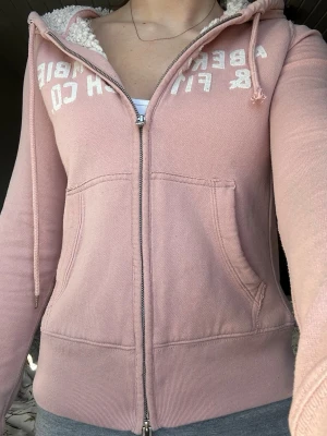Rosa vintage  hoodie från Abercrombie & Fitch - Mysig rosa hoodie med dragkedja från Abercrombie & Fitch. Stor vit logga på bröstet, två fickor fram och fluffigt teddyfoder i huvan. Långa ärmar och ribbade muddar. Perfekt för chill dagar och snygg till jeans eller tights. Den har en liten svart fläck på armen(bild 5) men enligt mig så tänker man inte så mycket på det