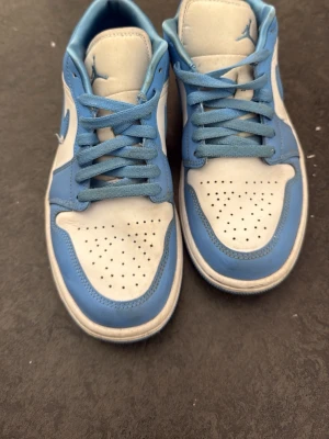 Jordan 1 Low UNC blå och vita sneakers - Snygga Jordan 1 Low sneakers, dom är använda 1 gång men det regnade ute första gången jag använde dom. Har ej använt dom efter då dom är förstora på mig.