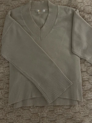 Beige stickad tröja från Gina Tricot - Beige stickad tröja från Gina Tricot med djup v-ringning och breda ribbade muddar vid ärmslut och nederkant. Tröjan har en avslappnad passform och är perfekt för lager-på-lager. Mjuk och skön att bära.