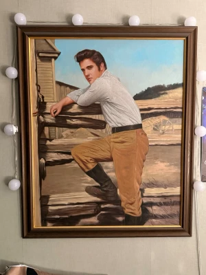 Tavla med Elvis Presley motiv i ram nyskick gjord för ca 40 år sen, sen har den gått i arv av min mamma! ETT MÅSTE FÖR ALLA ELVISFAN!!! - Säljer en stor tavla med motiv av Elvis Presley som lutar sig mot ett trästaket. Tavlan har en brun träram och bakgrunden visar en lantlig miljö med vagnhjul och fält. Elvis bär ljus skjorta, bruna byxor och svarta stövlar. Perfekt som dekor i hemmet. Mycket fint skick! Oljemålning 