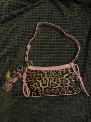 Leopardmönstrad handväska med rosa detaljer - Snygg handväska med leopardmönstrad textil och rosa konstläderdetaljer. Väskan har en axelrem i rosa, dekorativa knutar på sidorna och ett gulligt nyckelband med en glittrig hund. Perfekt för dig som gillar djurmönster och vill sticka ut.