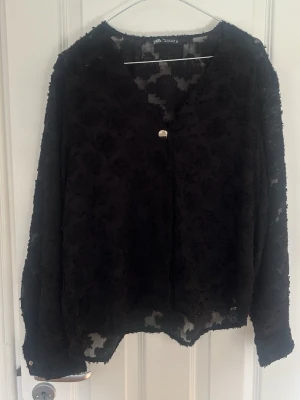 Svart blus med struktur från Zara - Snygg svart blus från Zara med långärmad design och v-ringning. Blusen har ett strukturerat, mönstrat tyg och en dekorativ knapp framtill. Materialet är lätt och något transparent, vilket ger en cool och trendig look.