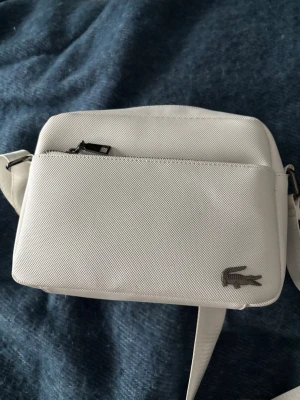 Vit axelväska från Lacoste - Snygg vit axelväska från Lacoste med justerbar rem och silverfärgad krokodillogga. Väskan har en stilren, rektangulär form och ett ytterfack med dragkedja. Tillverkad i slitstarkt tygmaterial som ger en sportig och modern känsla.