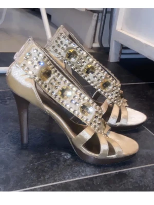 Guldiga klackskor - Snygga guldiga klackskor med öppen tå och glittrande strassdetaljer. Skorna har en elegant design med remmar och en bekväm klack. Perfekta för att ge en glamorös touch till din outfit. Enda defekten är undersidan, bild 3