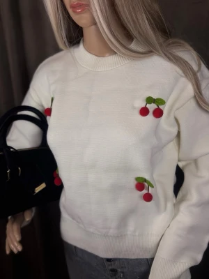 Vit stickad tröja med körsbärsdetaljer - Söt vit stickad tröja med broderade körsbär i rött och grönt på bröstet och sidan. Tröjan har rund halsringning, långa ärmar och ribbade muddar. Perfekt för dig som gillar lekfulla detaljer och vill ha en mysig men trendig look.