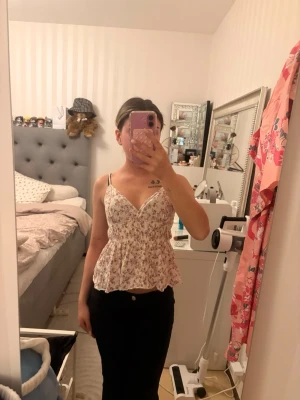 Blommig vit top - Söt blommig peplumtop med tunna axelband. Toppen är vit med rosa och beige blommönster, har en v-ringning och markerad midja som ger en feminin siluett. Perfekt för sommarens alla tillfällen.