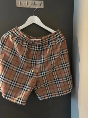 Rutigta shorts från Burberry - Snygga rutigta shorts från Burberry i klassiskt beige med svarta, vita och röda rutor. De har elastisk midja och är perfekta för sommaren. Ikonisk design som sticker ut och passar till många olika stilar.