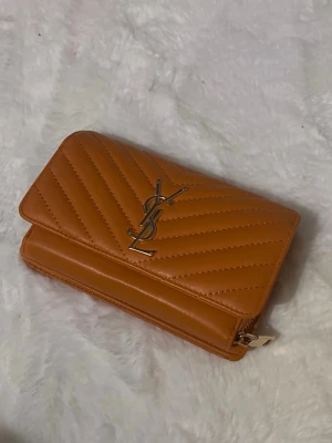 Brun YSL plånbok i läder - Säljer en snygg brun plånbok från YSL i quiltat läder med guldfärgad logga framtill. Plånboken har dragkedja på sidan och klassisk vikbar design. Perfekt för dig som gillar lyxiga accessoarer och vill ha något stilrent i väskan. 20cm lång 13hög