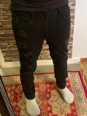Svarta ripped jeans med slim fit - Säljer ett par svarta jeans med slim fit och slitna detaljer på benen. Jeansen har klassisk femficksmodell och knappgylf. Snygg streetstil som passar perfekt till sneakers och t-shirt. Materialet känns mjukt och stretchigt för extra komfort.