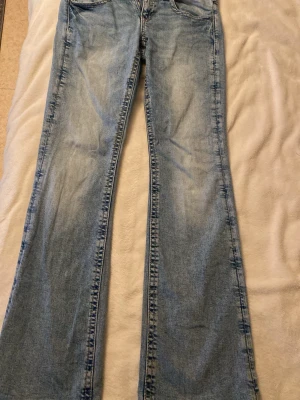 Ljusblå bootcut jeans - Säljer ett par ljusblå bootcut jeans med klassisk femficksdesign och normalhög midja. Jeansen har slitningar och tvättade detaljer för en avslappnad look. Perfekta för dig som gillar retrostil och vill ha ett par bekväma jeans i bomull.