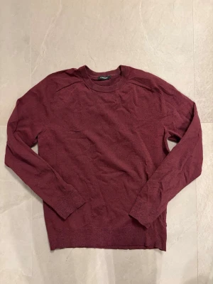Mörkröd stickad tröja från J. Lindeberg - Mörkröd stickad tröja från J. Lindeberg med rund halsringning och långa ärmar. Tröjan har en enkel och stilren design, perfekt för lager-på-lager och passar till många olika outfits.
