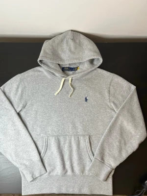 Grå hoodie från Polo Ralph Lauren - ÄKTA Polo Ralph Lauren hoodie i klassisk grå färg. Stilren design med broderad logga på bröstet och bekväm passform. Mjuk och skön kvalitet med huva och magficka – perfekt till både vardag och chill. Ett tidlöst plagg som aldrig går ur stil.