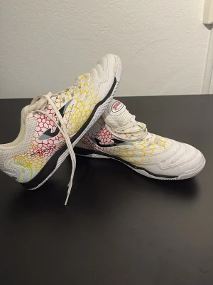 Vita Joma fotbollsskor med färgdetaljer - Säljer ett par vita fotbollsskor från Joma med coola mönster i gult, rött och grönt på sidorna. Skorna har låg profil, snörning och svart Joma-logga. Ovandelen är i syntetmaterial och sulan är anpassad för inomhusspel. Perfekta för dig som vill sticka ut på planen!