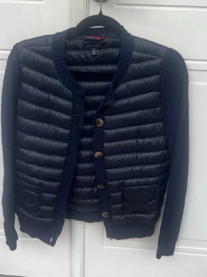 Tommy Hilfiger cardigan  - Snygg mörkblå cardiganjacka från Tommy Hilfiger med quiltade dunpaneler framtill och baktill. Stickade ärmar och kanter, rund halsringning och knäppning med träknappar. Två fickor framtill. Perfekt för lager-på-lager-stil.