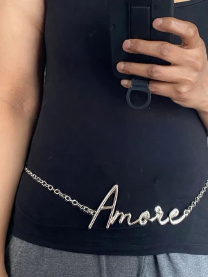 Silverfärgat kedjebälte med Amore-text - Säljer ett trendigt kedjebälte i silverfärgad metall med ordet 'Amore' i stor skrivstil framtill. Perfekt accessoar för att lyfta din outfit med en cool detalj. Justerbar längd och lätt att styla över klänning, kjol eller byxor.