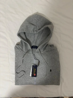 Grå hoodie från Polo Ralph Lauren - Klassisk grå hoodie från Polo Ralph Lauren med dragkedja och snörning i huvan. Ikonisk broderad logga på bröstet. Tillverkad i mjuk bomullsmix för en skön och avslappnad stil.